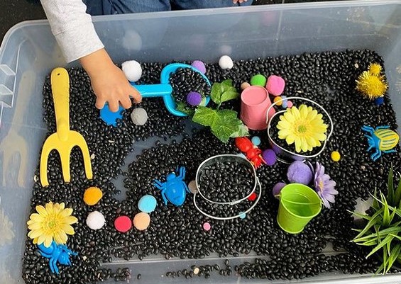 Spring sensory bin with black beans, pots, garden tools, flowers, fake grass.