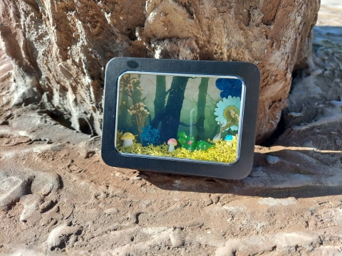 A small black tin rests on a tree, showcasing its content; Three miniature mushrooms and one alligator sitting atop fake moss.