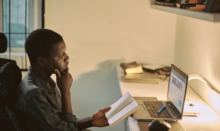 Male teen reading a book at a desk with an open laptop